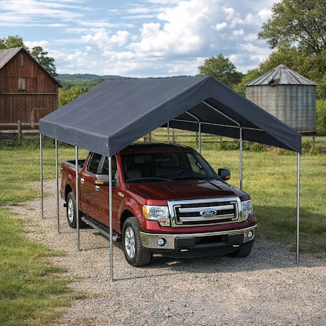 Red truck under a gray carport in a rural setting with a barn and silo in the background.